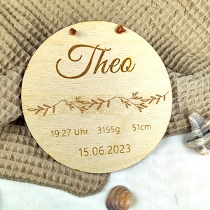 May include: A round wooden plaque with the name "Theo" engraved in cursive script. It also includes the date "15.06.2023", time "19:27 Uhr", weight "3155g", and length "51cm".