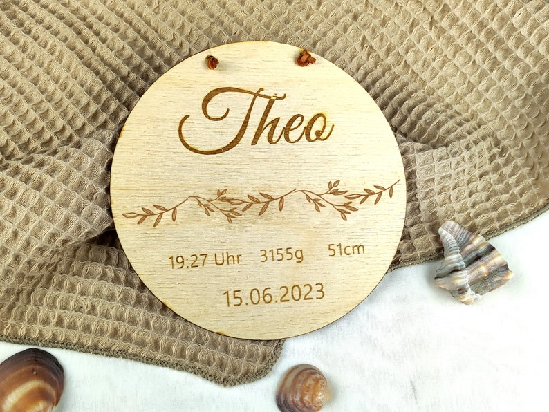May include: A round wooden plaque with the name "Theo" engraved in cursive script. It also includes the date "15.06.2023", time "19:27 Uhr", weight "3155g", and length "51cm".