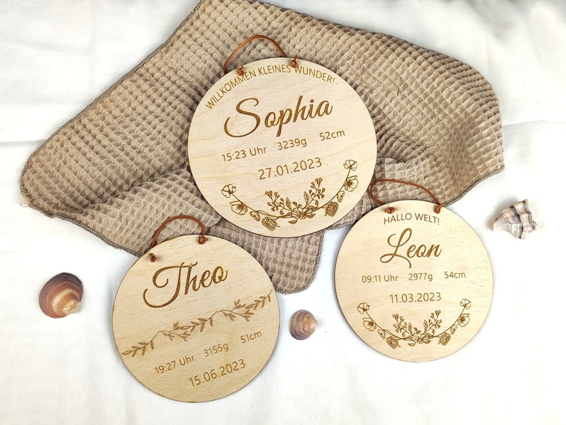 May include: Three wooden round plaques with engraved text and floral designs. The plaques feature the names Sophia, Theo, and Leon, along with birth details including date, time, weight, and length. The plaques are on a beige textured surface.