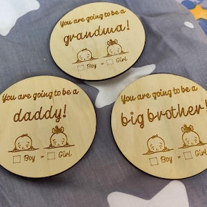 May include: Three wooden plaques with the text "You are going to be a grandma!"  "You are going to be a daddy!" and "You are going to be a big brother!" Each plaque has a cartoon image of a baby peeking over a line with a checkbox for "Boy" or "Girl".