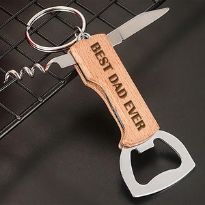Könnte beinhalten: Ein Holzschlüsselanhänger mit einem Flaschenöffner, einem Korkenzieher und einem kleinen Messer. Der Schlüsselanhänger ist mit dem Text "Best Dad Ever" graviert.