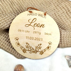 May include: A round wooden sign with the text "Hallo Welt!" and "Leon" in cursive. The sign also includes the date "11.03.2023", the time "09:11 Uhr", and the measurements "2977g" and "54cm". The sign is decorated with a floral design.