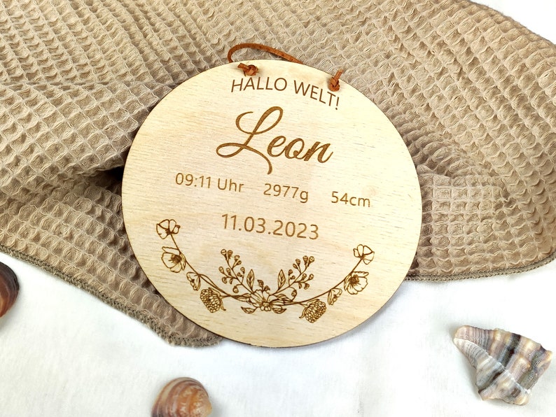 May include: A round wooden sign with the text "Hallo Welt!" and "Leon" in cursive. The sign also includes the date "11.03.2023", the time "09:11 Uhr", and the measurements "2977g" and "54cm". The sign is decorated with a floral design.