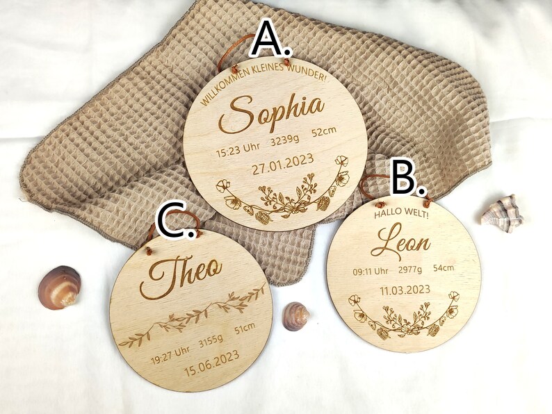 May include: Three wooden plaques with engraved text and floral designs. The plaques are labeled A, B, and C. Plaque A reads "Willkommen kleines Wunder! Sophia 15:23 Uhr 3239g 52cm 27.01.2023". Plaque B reads "Hallo Welt! Leon 09:11 Uhr 2977g 54cm 11.03.2023". Plaque C reads "Theo 19:27 Uhr 3155g 51cm 15.06.2023".
