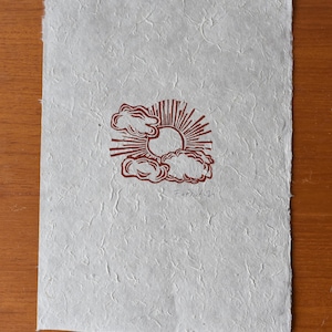 May include: A handmade paper print featuring a brown sun and cloud design. The artwork has a textured surface and a deckled edge. The artist's signature and the year '21 are visible in the lower right corner.