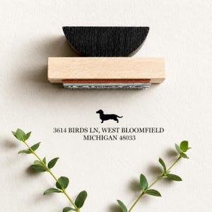 May include: A wooden stamp featuring a dachshund silhouette and the address 3614 BIRDS LN, WEST BLOOMFIELD, MICHIGAN 48033. The stamp has a black handle and a light wood base. Green leaves are at the bottom.