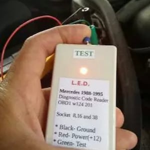 Puede incluir: Un lector de código de diagnóstico blanco para vehículos Mercedes de 1988 a 1995. El dispositivo tiene una luz LED roja y texto que dice "TEST", "L.E.D.", "Mercedes 1988-1995", "Diagnostic Code Reader", "OBD1 w124 201", "Socket 8, 16 and 38", "Black-Ground", "Red-Power(+12)" y "Green-Test".
