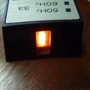 May include: A black box with a small orange light shining through a rectangular opening. The box has a silver label with the text "60Hz 33 60Hz 09".