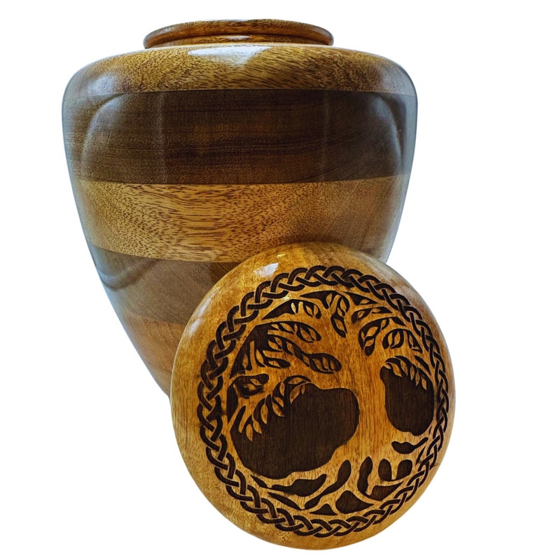 Pu&ograve; includere: Un'urna di legno con coperchio rimovibile. L'urna &egrave; realizzata con fasce alternate di legno chiaro e scuro. Il coperchio presenta un'intagliatura dettagliata di un albero all'interno di un bordo a nodo celtico.