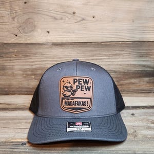 Puede incluir: Gorra de camionero gris y negra con un parche que presenta un pato de dibujos animados con gafas de sol sosteniendo una pistola, y las palabras "PEW PEW MADAFKAS!". La gorra tiene una visera curva y una parte trasera de malla.