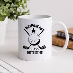 May include: White ceramic coffee mug with a black graphic of crossed golf clubs and a golf ball. The text reads "Weapons of Grass Destruction".