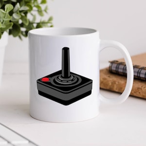 May include: White ceramic mug with a black and white illustration of a vintage joystick video game controller. The joystick has a red button.