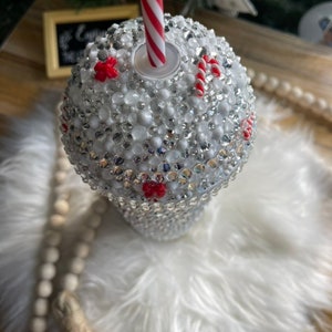 May include: White tumbler with a red and white striped straw. The tumbler is covered in rhinestones and has red and white candy cane accents.
