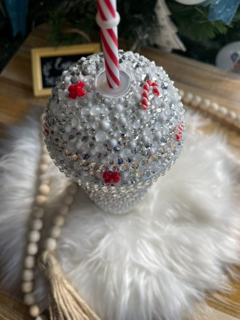 May include: White tumbler with a red and white striped straw. The tumbler is covered in rhinestones and has red and white candy cane accents.