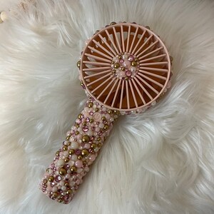 Puede incluir: Un ventilador de mano rosa con un mango de cuentas blancas y doradas. El ventilador tiene una carcasa de pl&aacute;stico rosa y una h&eacute;lice de pl&aacute;stico blanca.