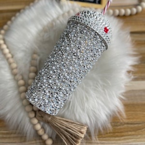 May include: A white tumbler with a silver glitter design and a red and white striped straw. The tumbler has a clear lid with a silver glitter design. The tumbler is sitting on a white fluffy surface.