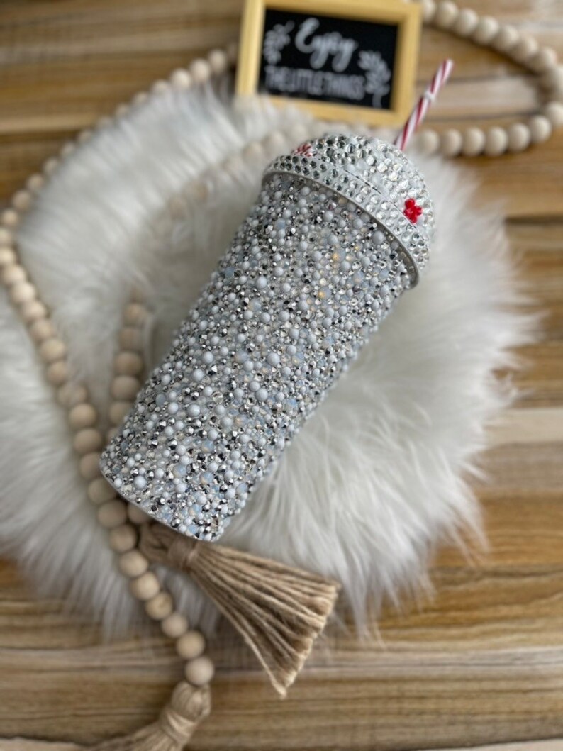 May include: A white tumbler with a silver glitter design and a red and white striped straw. The tumbler has a clear lid with a silver glitter design. The tumbler is sitting on a white fluffy surface.