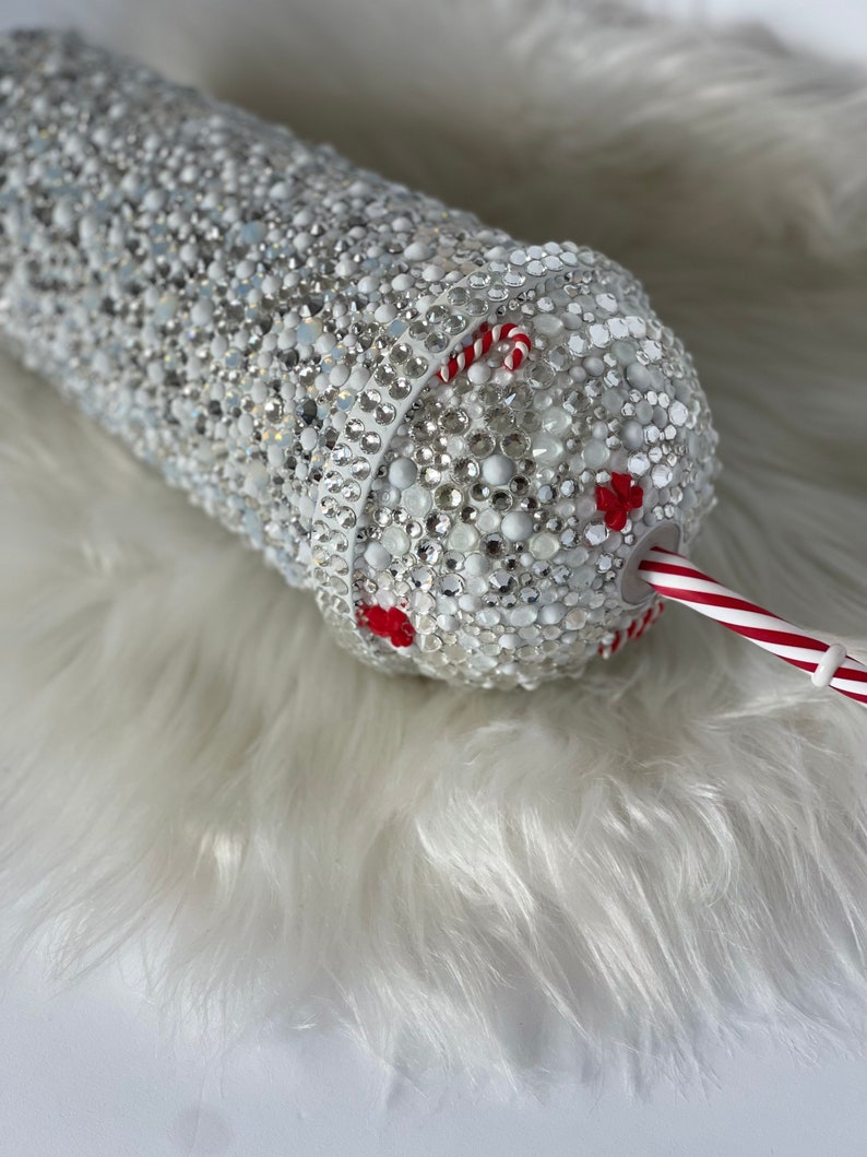 May include: A white tumbler covered in rhinestones with a red and white candy cane straw.