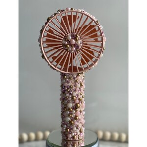 Puede incluir: Un ventilador de mano rosa con un dise&ntilde;o de cuentas blancas y doradas. El ventilador tiene una cuchilla redonda rosa y un bot&oacute;n blanco en el mango.