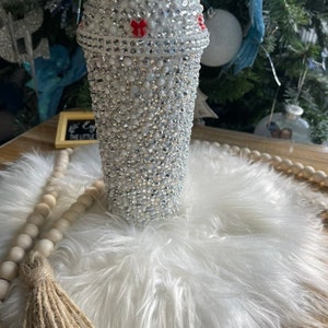 May include: A white tumbler with a red and white striped straw. The tumbler is covered in rhinestones and has a red bow on the lid. The tumbler is sitting on a white fluffy surface.