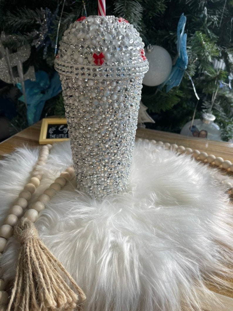 May include: A white tumbler with a red and white striped straw. The tumbler is covered in rhinestones and has a red bow on the lid. The tumbler is sitting on a white fluffy surface.