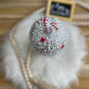 May include: A white wine glass with a red and white candy cane design and a silver straw. The glass is covered in rhinestones.