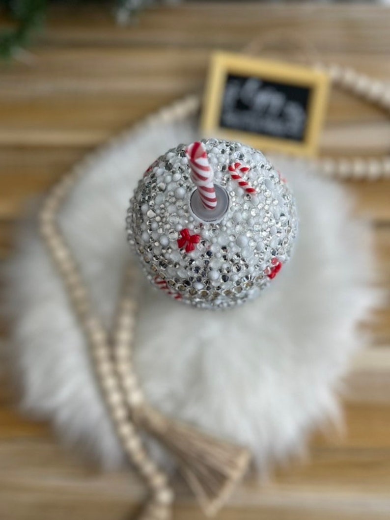 May include: A white wine glass with a red and white candy cane design and a silver straw. The glass is covered in rhinestones.