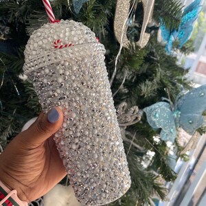 May include: A white tumbler with a red and white candy cane design. The tumbler is covered in rhinestones and has a clear plastic straw.