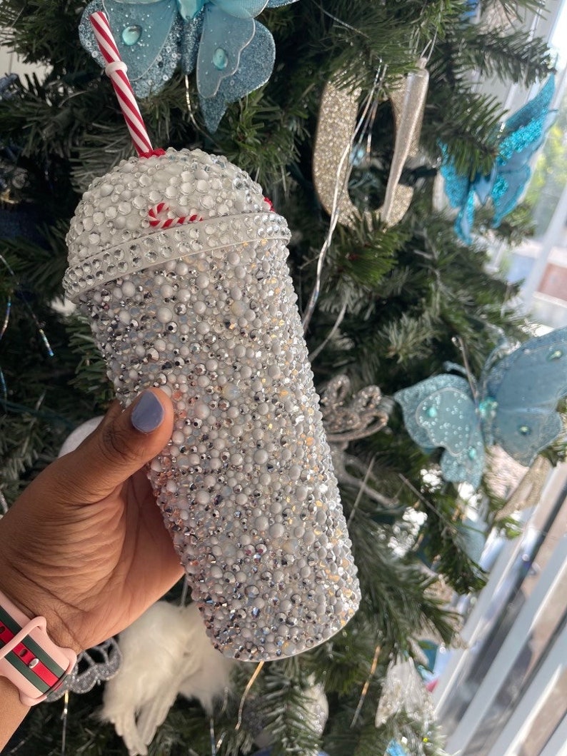 May include: A white tumbler with a red and white candy cane design. The tumbler is covered in rhinestones and has a clear plastic straw.