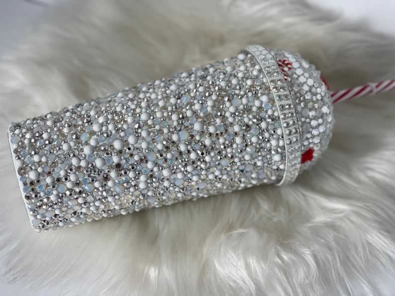 May include: A white tumbler covered in silver and white rhinestones with a red and white striped straw.