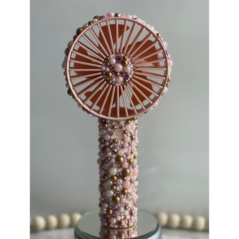 Puede incluir: Un ventilador de mano rosa con un dise&ntilde;o de cuentas blancas y doradas. El ventilador tiene una cuchilla redonda rosa y un bot&oacute;n blanco en el mango.