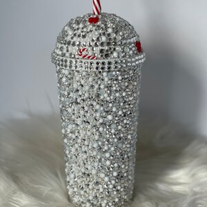 May include: A white tumbler with a red and white striped straw. The tumbler is covered in white and silver rhinestones. There are two red hearts on the top of the tumbler.
