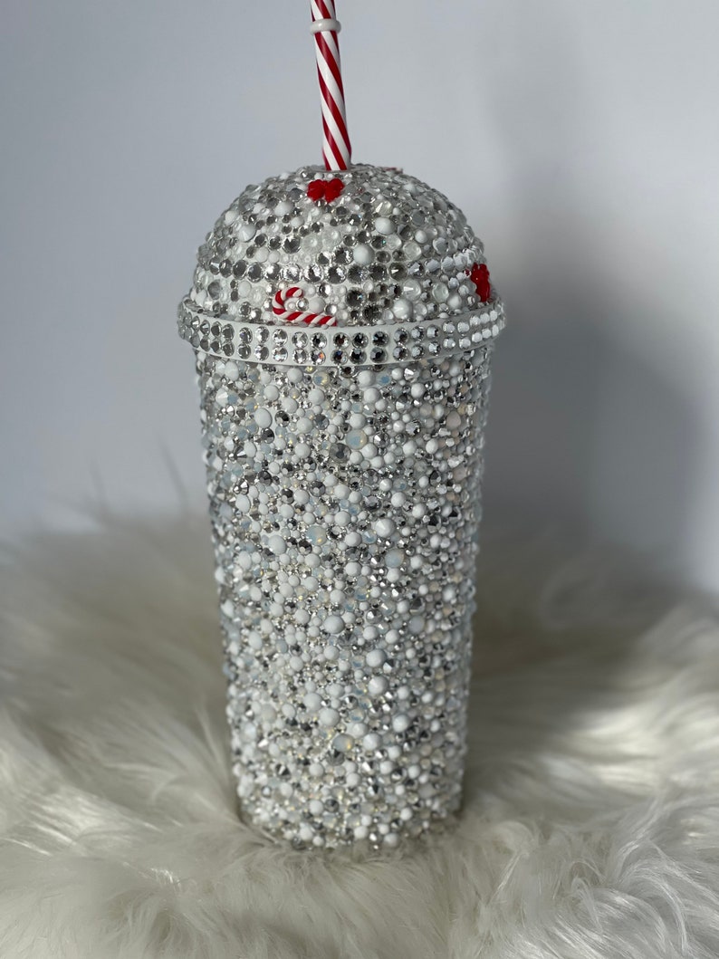 May include: A white tumbler with a red and white striped straw. The tumbler is covered in white and silver rhinestones. There are two red hearts on the top of the tumbler.