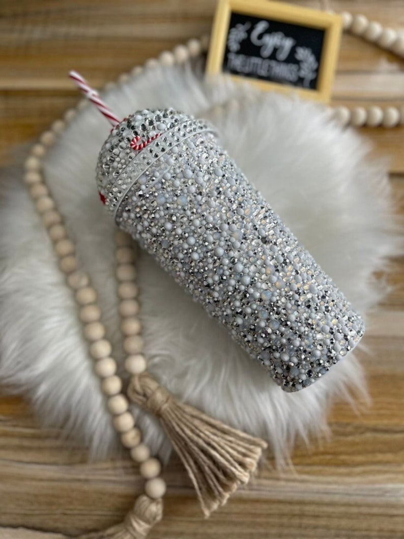 May include: A white tumbler with a red and white striped straw. The tumbler is covered in rhinestones and has a sparkly finish.