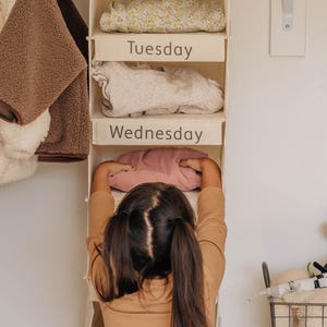 May include: A white fabric hanging organizer with six compartments labeled "Monday", "Tuesday", "Wednesday", "Thursday", "Friday", and "Saturday". A young person is putting clothes into the organizer.
