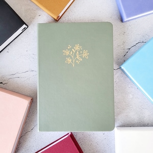 Op de afbeelding: Een saliegroen notitieboek met een gouden bloemenontwerp in het midden. Het is omringd door andere kleurrijke notitieboeken in goud, zwart, roze, blauw en paars. De notitieboeken liggen op een gestructureerd, lichtgrijs oppervlak.