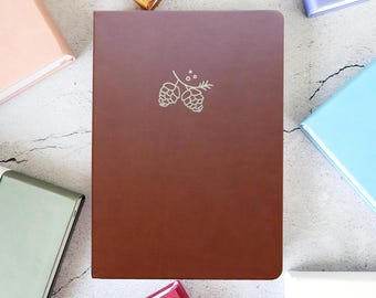 May include: A brown journal with a silver floral design is centered in the image, surrounded by other journals in various colors. The cover of the journal has a debossed floral design. The other journals are in shades of pink, gold, blue, and green.