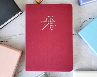 May include: A red hardcover journal with a white design of a wand and stars on the front cover.