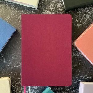 Op de afbeelding: Een bordeauxrood notitieboek met een wit lint als bladwijzer. Het notitieboek is gesloten en ligt plat op een donker, getextureerd oppervlak.