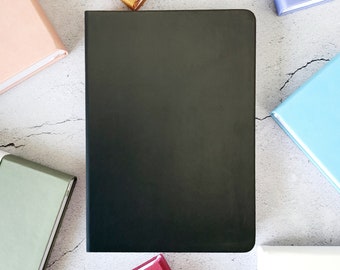May include: A black notebook is the focal point, surrounded by other colorful notebooks in shades of pink, gold, purple, blue, red, and green. The notebooks are arranged on a textured, light gray surface.