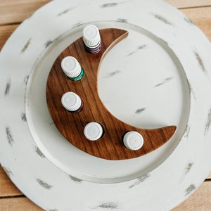 Può includere: Un porta oli essenziali a forma di mezzaluna in legno con cinque piccole bottiglie. Il supporto è posto su un grande piatto bianco dall'aspetto invecchiato. Le bottiglie hanno tappi bianchi e contengono liquidi di vari colori.