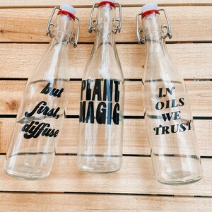 May include: Three clear glass bottles with black lettering on a wood background. The bottles have swing top lids. The text on the bottles reads "but first, diffuse", "plant magic", and "in oils we trust".