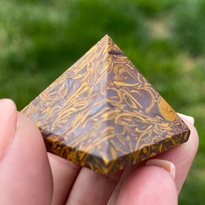 May include: A pyramid-shaped decorative stone with a brown base and yellow, swirled patterns. The pyramid is held in a hand against a blurred green background.