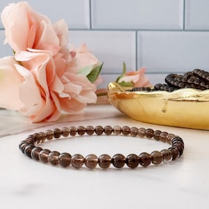 May include: A brown gemstone bracelet made with small, round beads. The bracelet is lying on a white surface with a pink flower in the background.