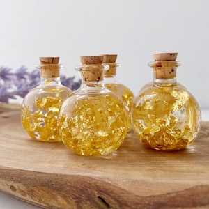 May include: Five small, clear glass bottles with cork stoppers, filled with gold leaf. The bottles sit on a wooden surface, with a blurred background of lavender and a white wall.