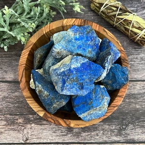 May include: A wooden bowl filled with vibrant blue lapis lazuli stones. A bundle of sage and greenery are also present. The stones have a deep blue color with flecks of gold and white. The bowl is made of wood with a natural grain.