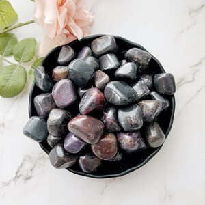May include: A black bowl filled with tumbled purple and black ruby stones. The stones are smooth and polished.