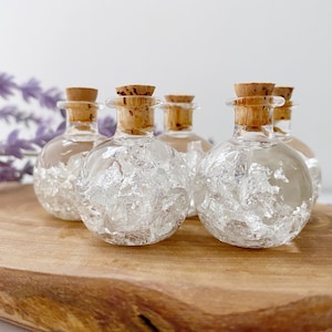 May include: Five small glass bottles with cork stoppers filled with silver flakes. The bottles are arranged on a wooden surface.