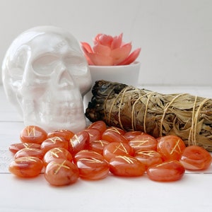 May include: A white ceramic skull, orange rune stones with gold markings, a bundle of sage, and a small succulent in a white pot. The rune stones are scattered in front of the other objects.