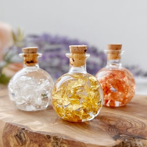 May include: Three small glass bottles with cork stoppers, each filled with a different material. The bottle on the left is filled with clear crystals, the middle bottle is filled with gold flakes, and the bottle on the right is filled with orange flakes.